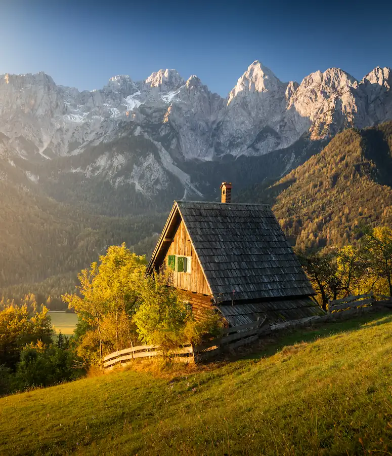 A rustic mountain hut in Srednji Vrh, Slovenia, overlooking a deep valley and the dramatic, sun-drenched peaks of the Julian Alps.