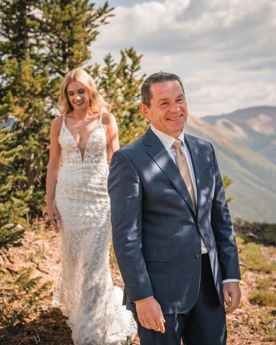 Clemmie approaches a smiling man in a blue suit from behind on a woodland path.