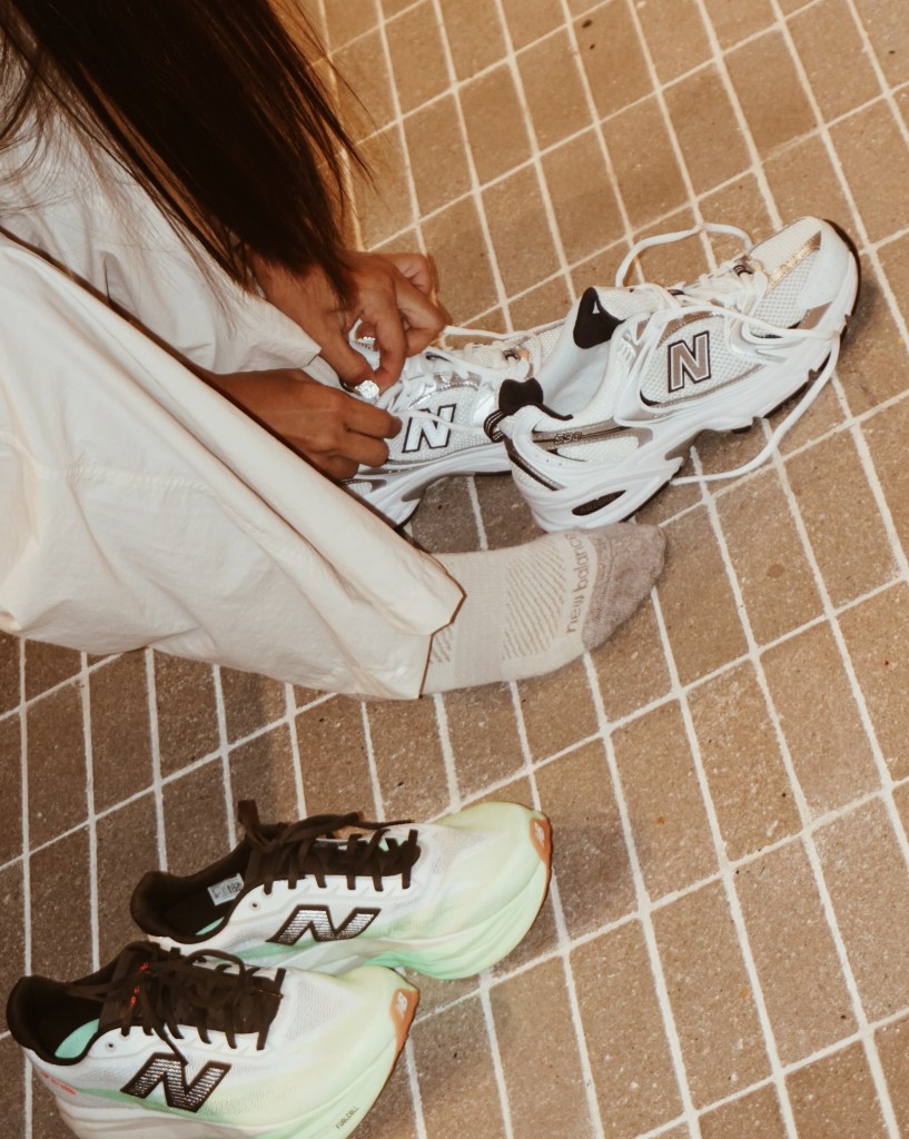 A person sitting on the floor, lacing up a pair of New Balance running shoes, with another pair of running shoes nearby, on a tiled surface.