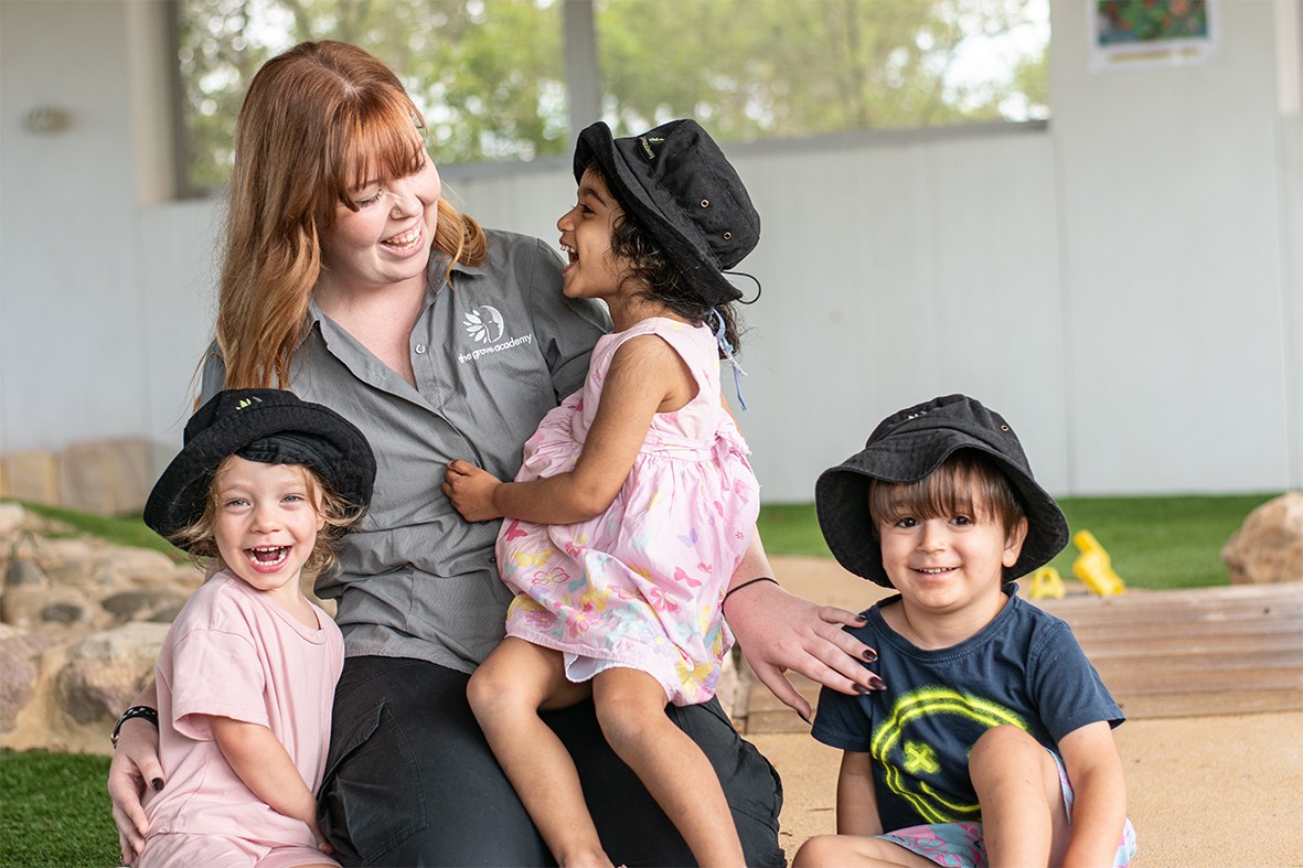Smiling young woman with long hair standing against a dark green background, holding a finger to her chin.