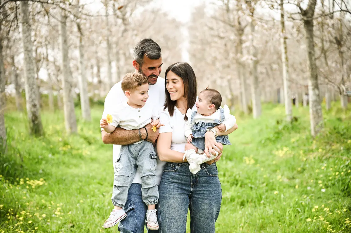 פורטרט משפחתי מלא חום בלב חורשה פורחת. זוג הורים מחייכים ומביטים בילדיהם הקטנים באווירה אביבית ונינוחה תחת אור טבעי, מאת Shiriz Photography הצלמת שירי עציץ.