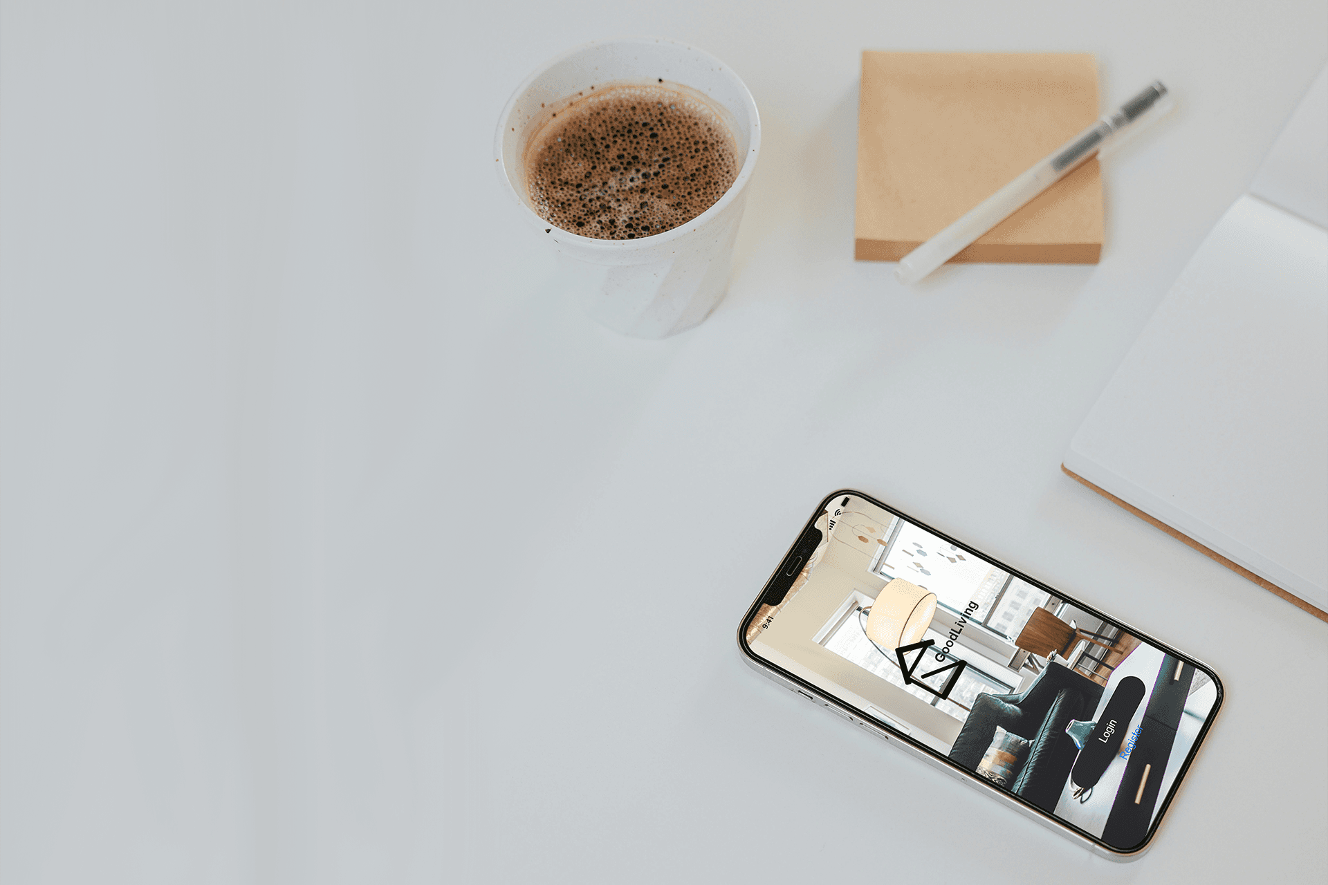 Eine weiße Tischoberfläche mit einem Smartphone, das die "GoodLiving"-App-Startseite zeigt, einer Tasse Kaffee, einem Notizblock, einem Stift und Haftnotizen. Das Telefon liegt im unteren rechten Bereich.