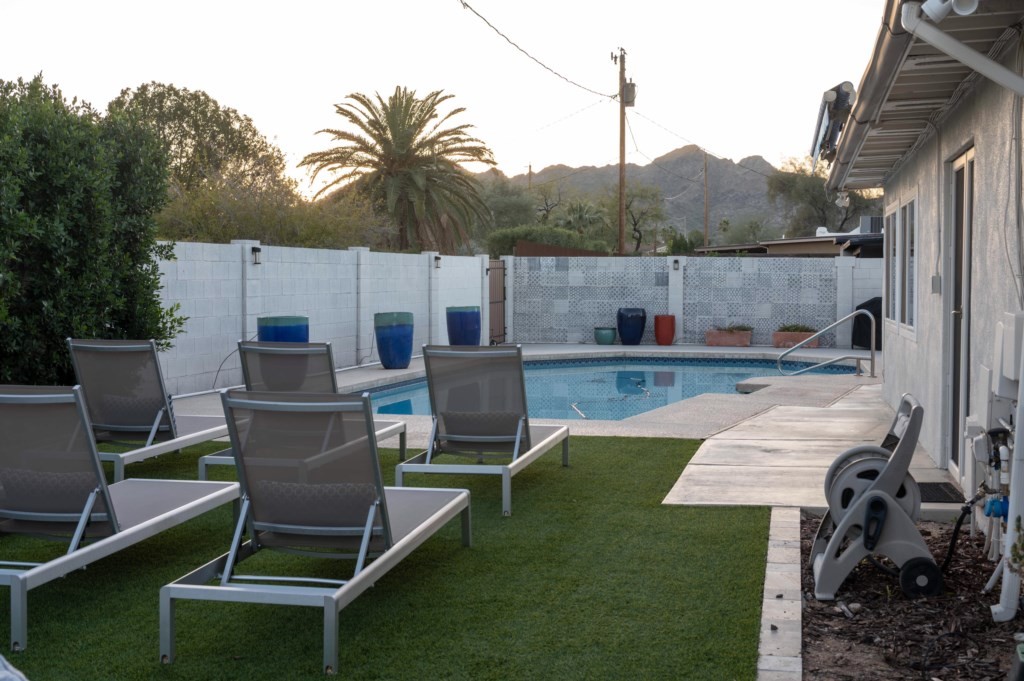 A serene backyard view of an affordable luxury vacation rental house, showcasing a sparkling swimming pool surrounded by sleek metal loungers, with desert landscaping and mountains visible in the distance.