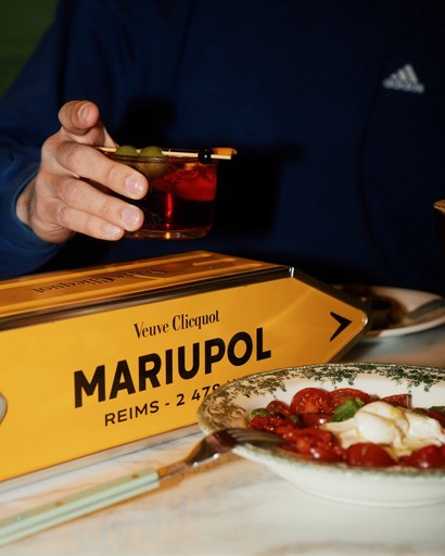 A person holds a cocktail garnished with green olives and a cherry next to a yellow Veuve Clicquot box labeled "Mariupol Reims - 247," with a plate of tomato and mozzarella salad in the foreground.
