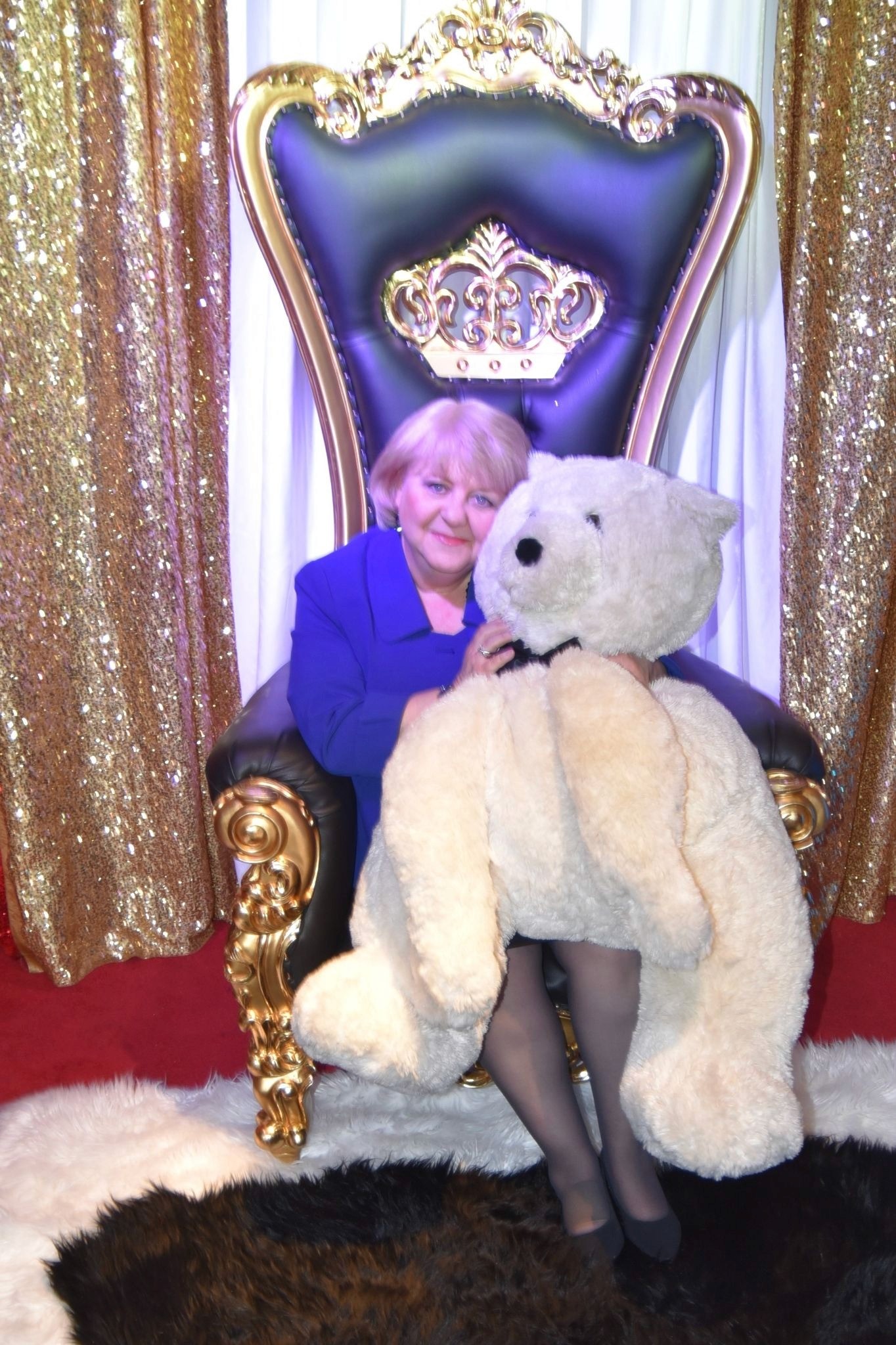 A person in a blue outfit sits on an ornate, gold-trimmed throne chair, hugging a large white teddy bear. The backdrop has glittering gold curtains, creating a festive atmosphere.