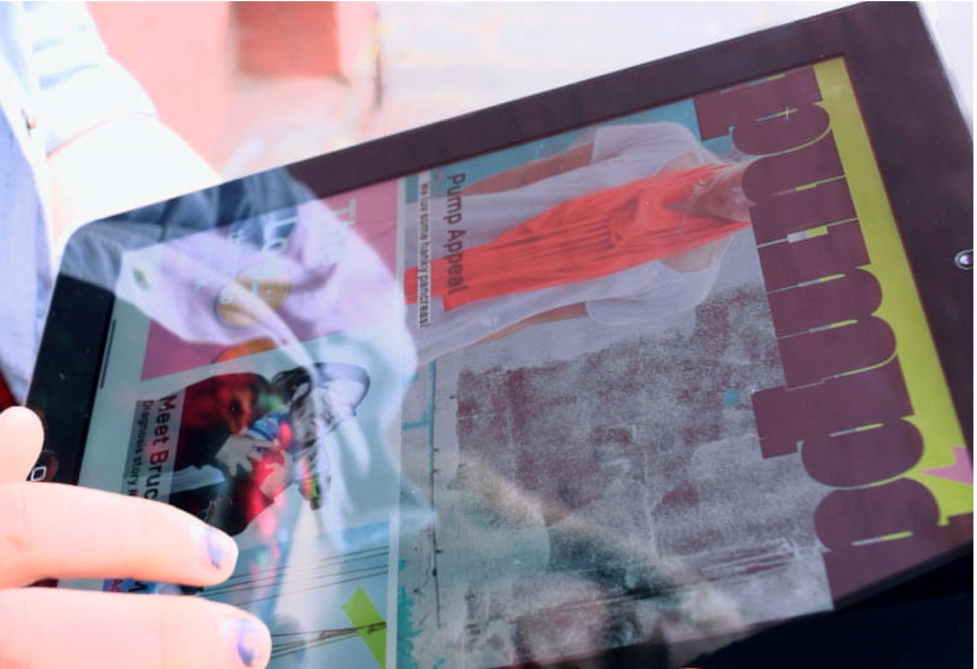 A hand with blue nail polish holds a tablet displaying a magazine cover. The cover features a woman in an orange top against a light background, with text including “Pimp Appeal” and other smaller text. The word “Pimp” appears in large brown letters with a yellow-green gradient along the right side. The magazine cover is partially obscured by the hand and tablet.