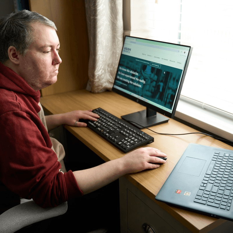 An image of a man using a light and magnifying tool to read a book, whilst sat on a couch.
