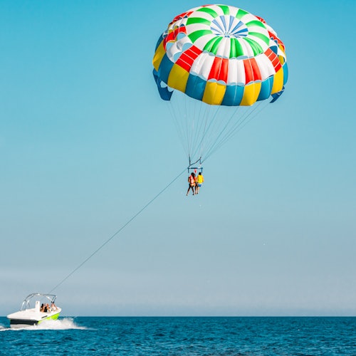 Two people parasailing over the ocean, towed by a white boat under a clear blue sky with a colorful parachute.