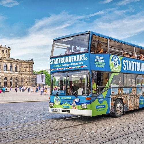 Kaksikerroksinen kiertoajelubussi, jossa lukee "STADTRUNDFAHRT", ajaa historiallisen rakennuksen edessä mukulakiviplazalla.