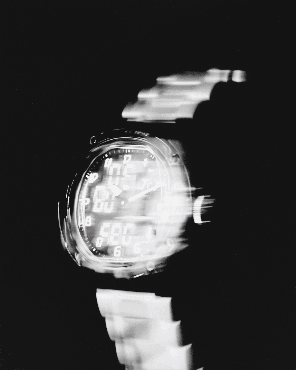 Black-and-white motion blur of a wristwatch with a metallic strap, blending analog and digital elements against a dark background.
