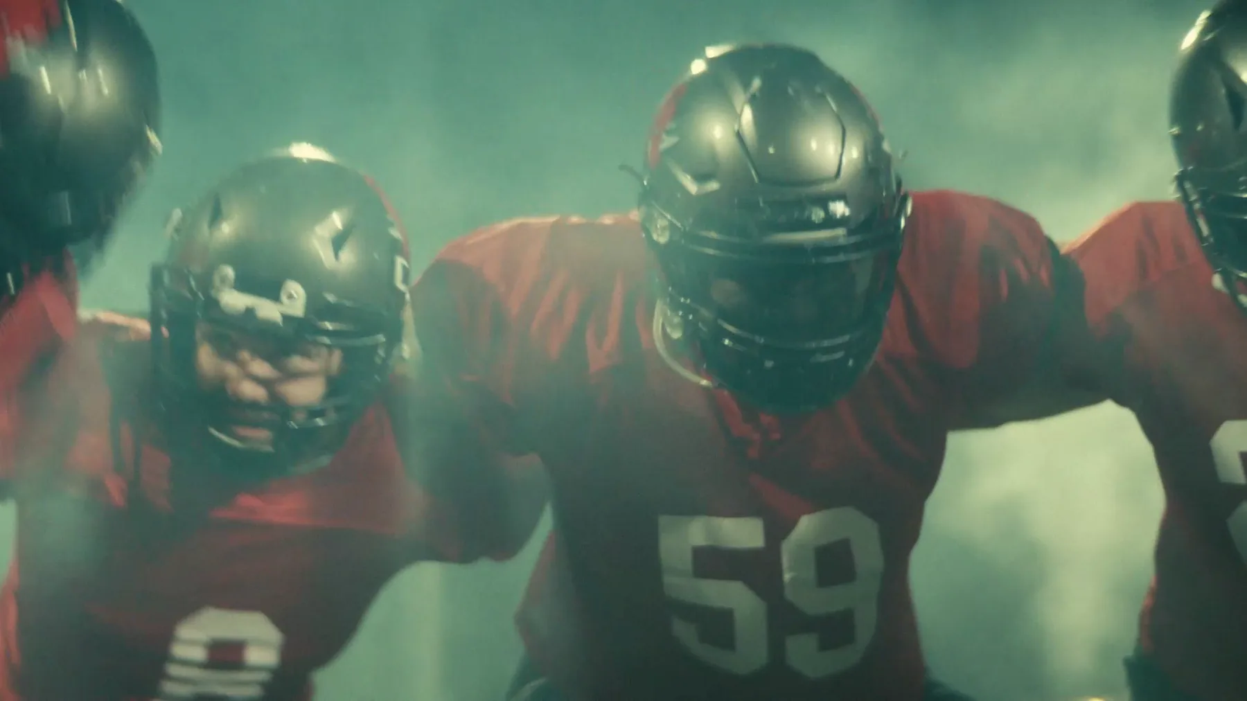 Three football players in red jerseys and dark helmets stand shoulder to shoulder in a huddle, backlit by green-tinted smoke.