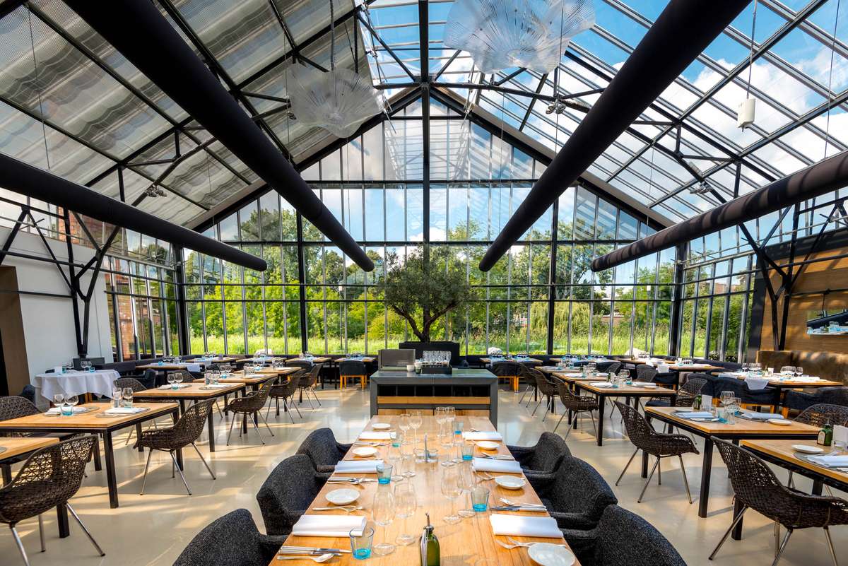 A glass-roofed conservatory restaurant with long tables set for dining and a large green tree in the center.