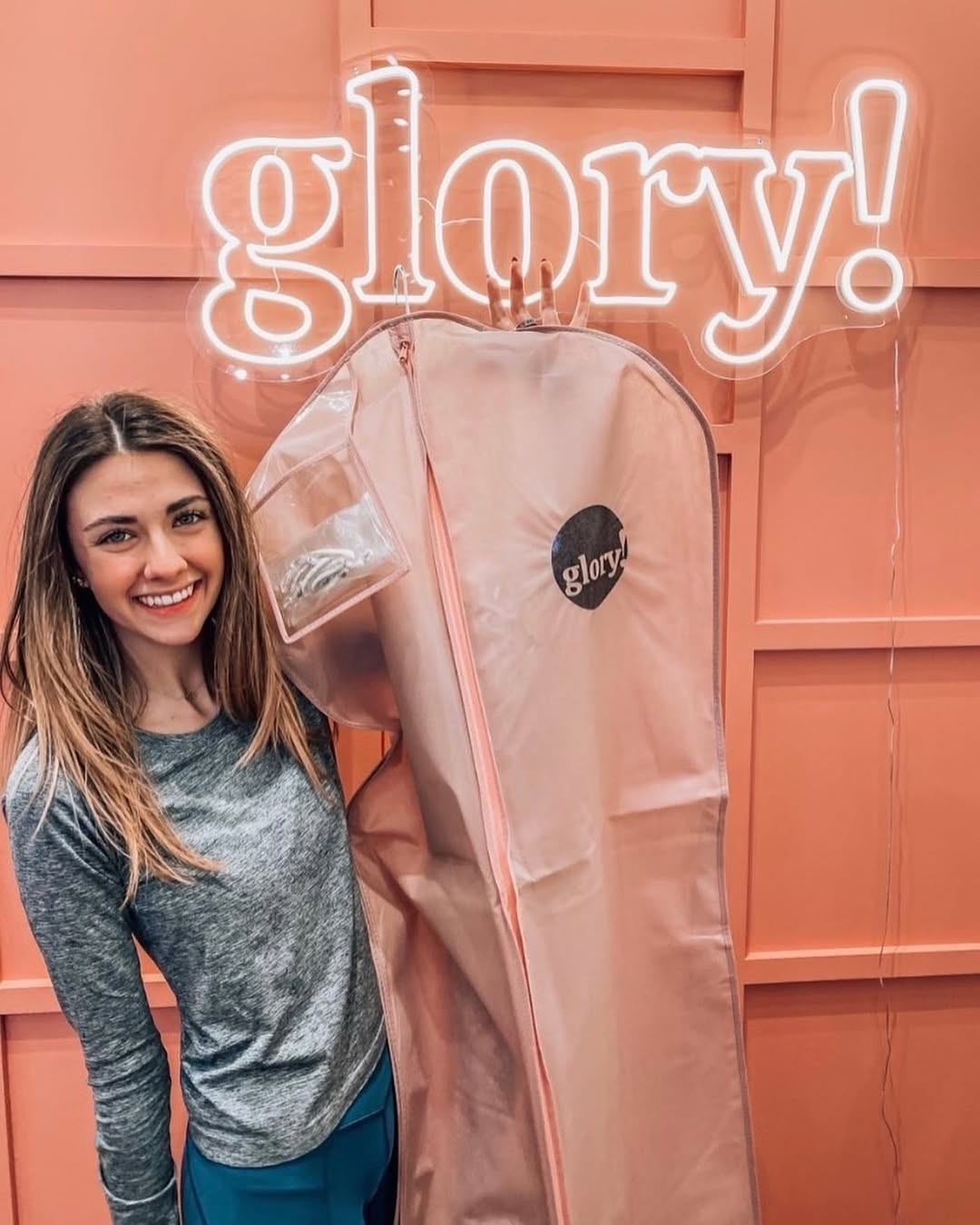 Woman standing beside a garment bag in front of a peach wall with an illuminated “glory!” neon sign.
