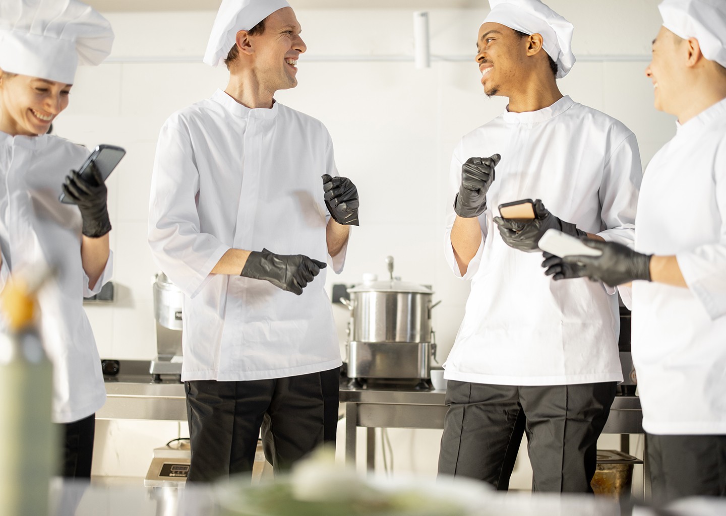 Sullo sfondo di una cucina il personale di un ristorante si scambia allegramente commenti mostrandosi a vicenda lo schermo degli smartphone