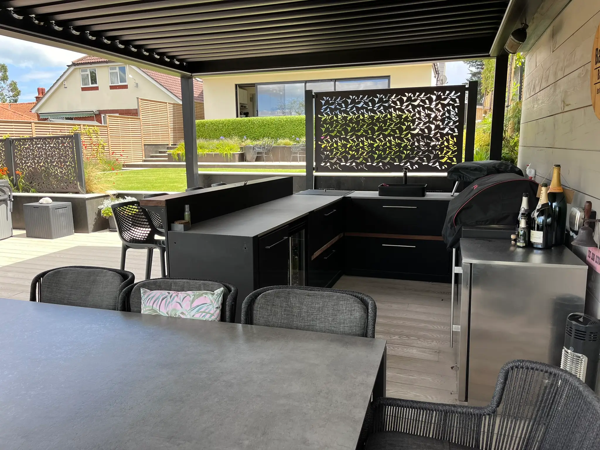 Outdoor dining area with sleek black tables and chairs, shaded by a canopy, surrounded by greenery.