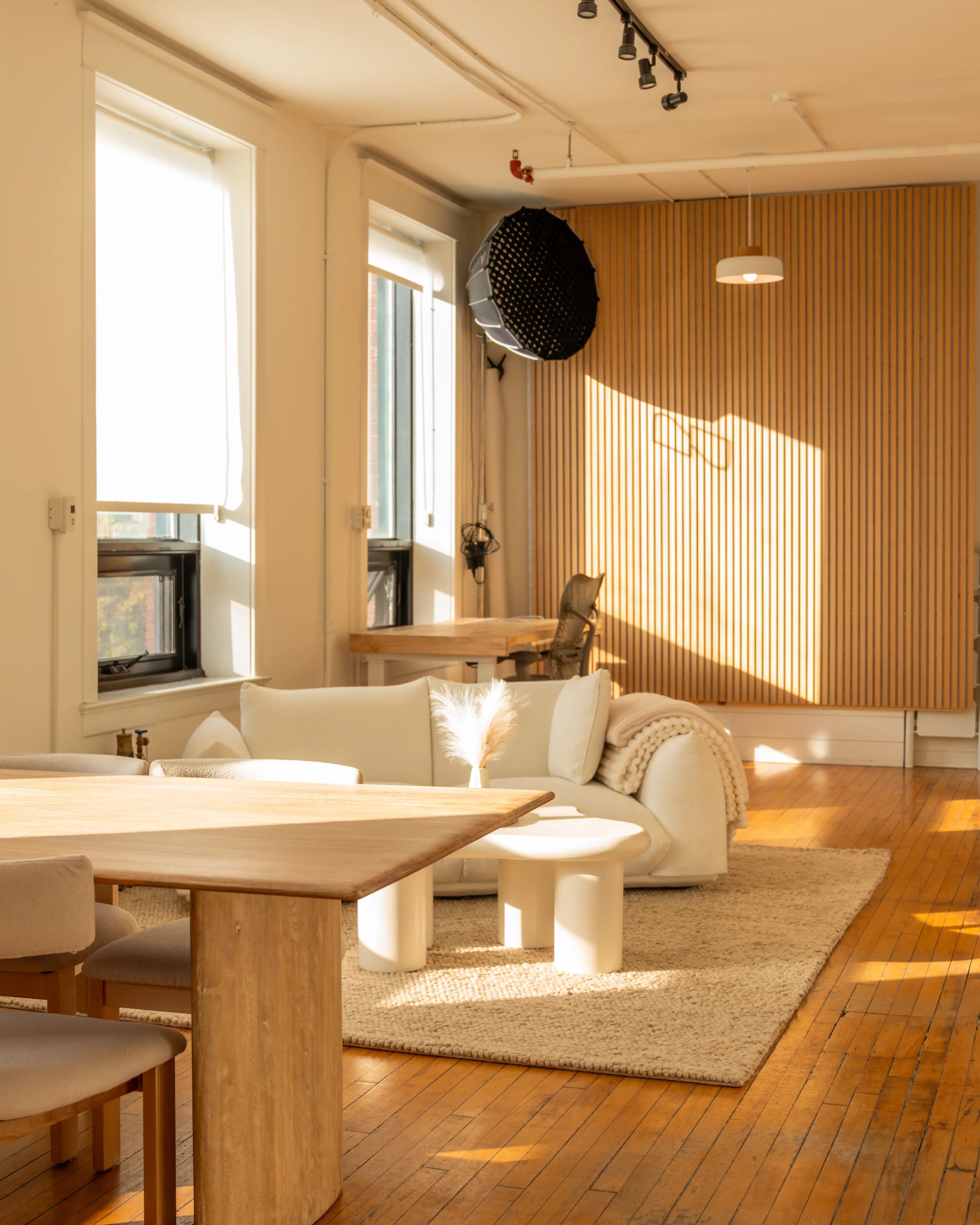 Node Studio’s open-concept layout with warm hardwood floors, natural light pouring through large windows, a cozy white couch on a textured rug, and a slatted wood accent wall creating a bright and inviting atmosphere