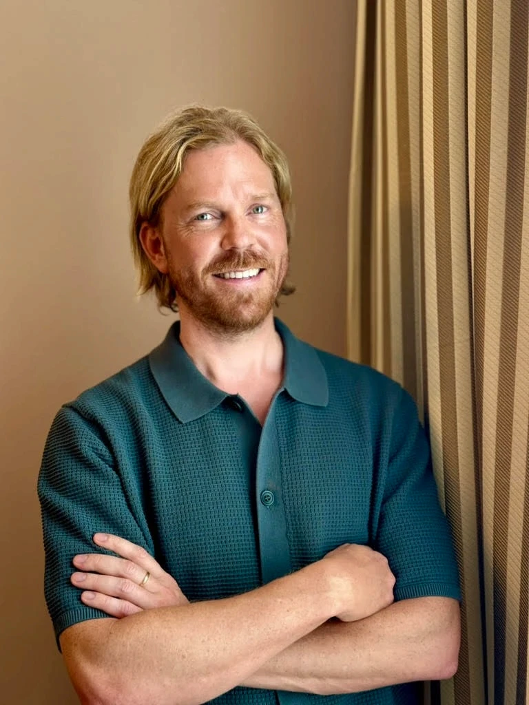 Smiling blond man with a beard and teal polo shirt. Leaning against a wall with striped curtain.