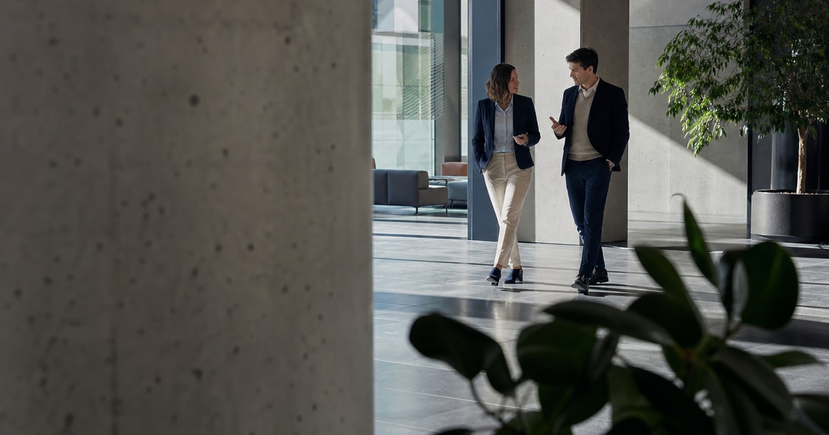 Two advisors walk through a sunlit modern office building foyer, framed past a concrete column. They are engaged in quiet conversation about market research. · K2 Advocacy