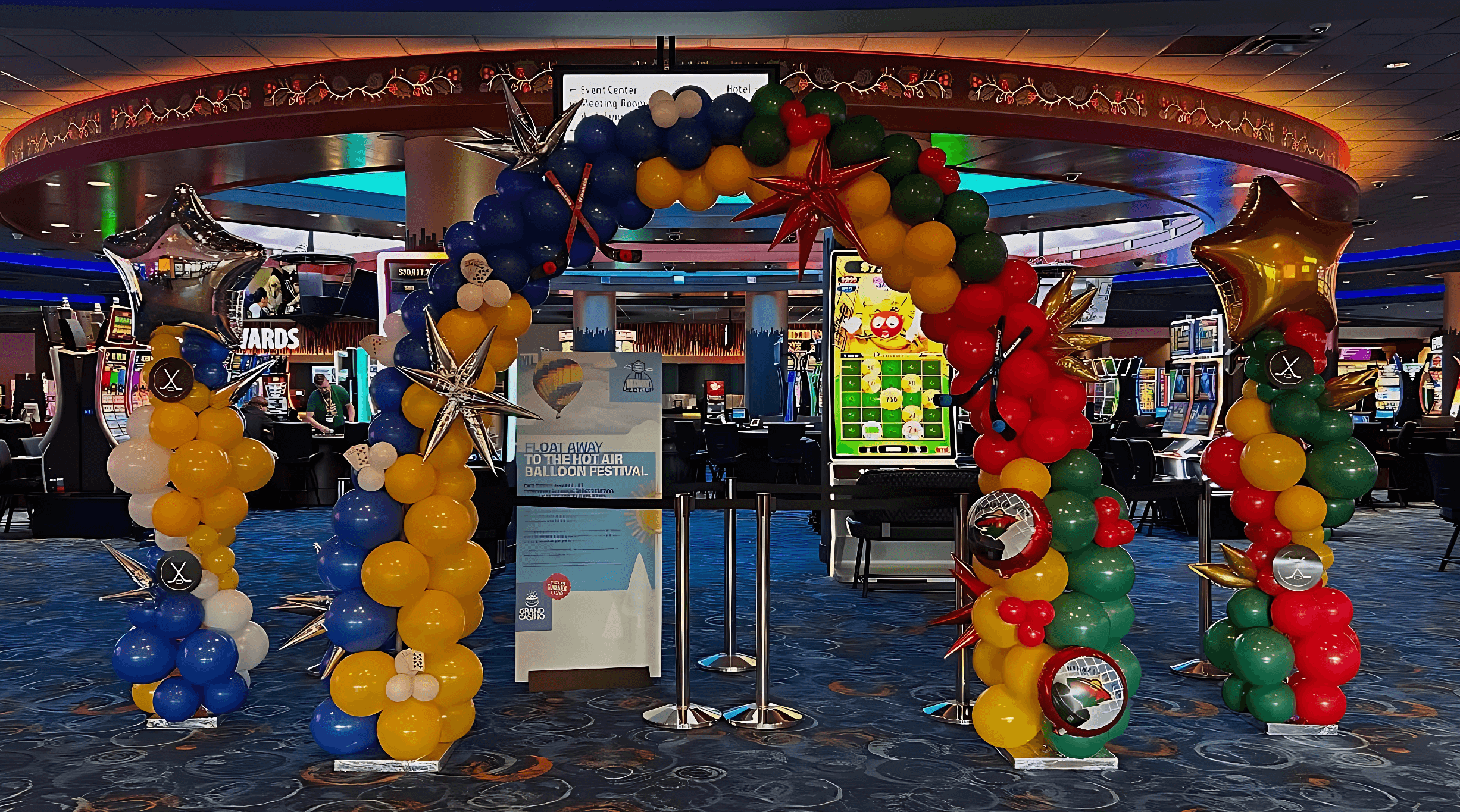 A vibrant balloon arch in blue, yellow, red, and green stands in a casino entrance, accented by gold stars and displayed in front of gaming machines.