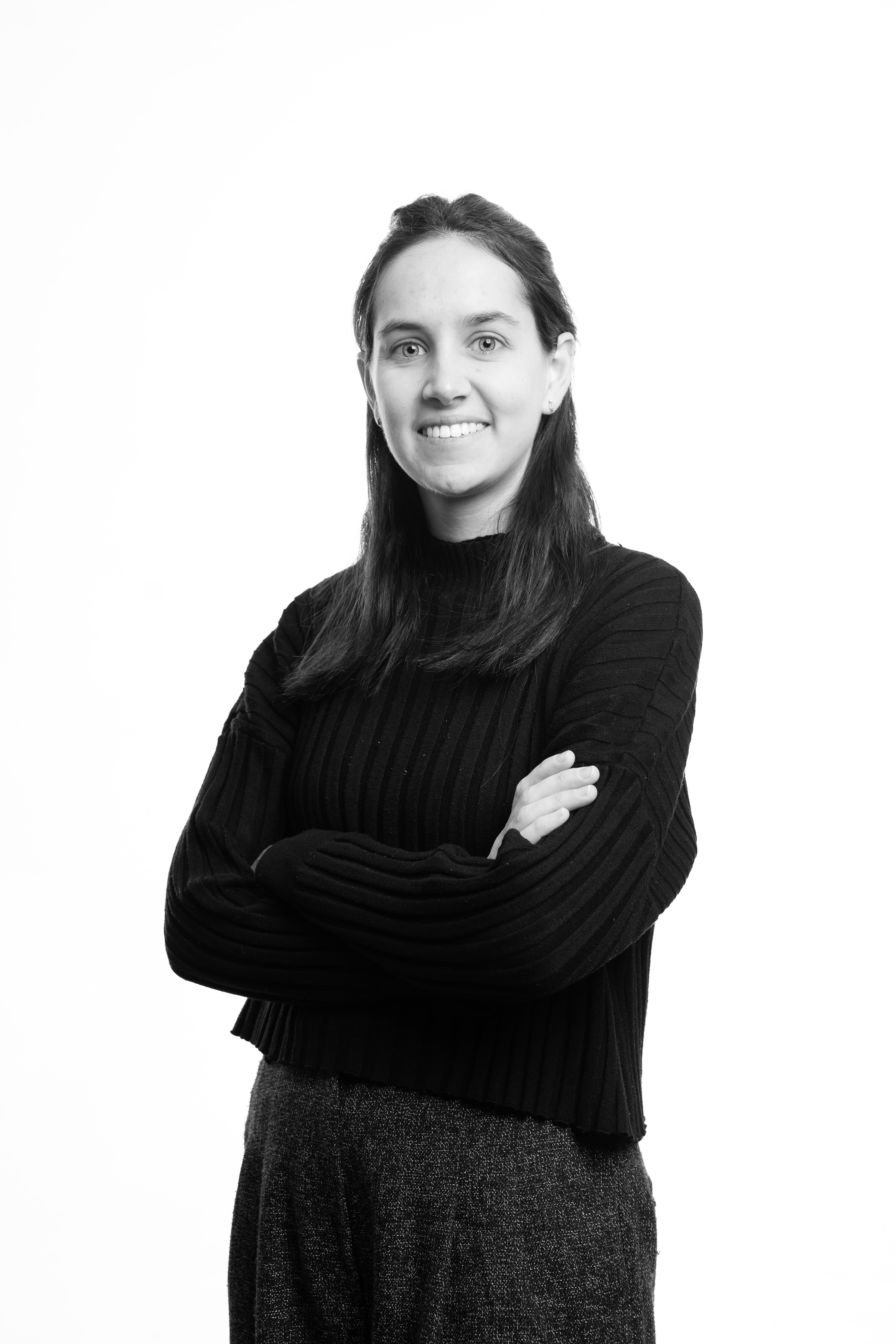 Black and white photo of a confident person with long hair, wearing a textured sweater. They are smiling with arms crossed against a plain background.