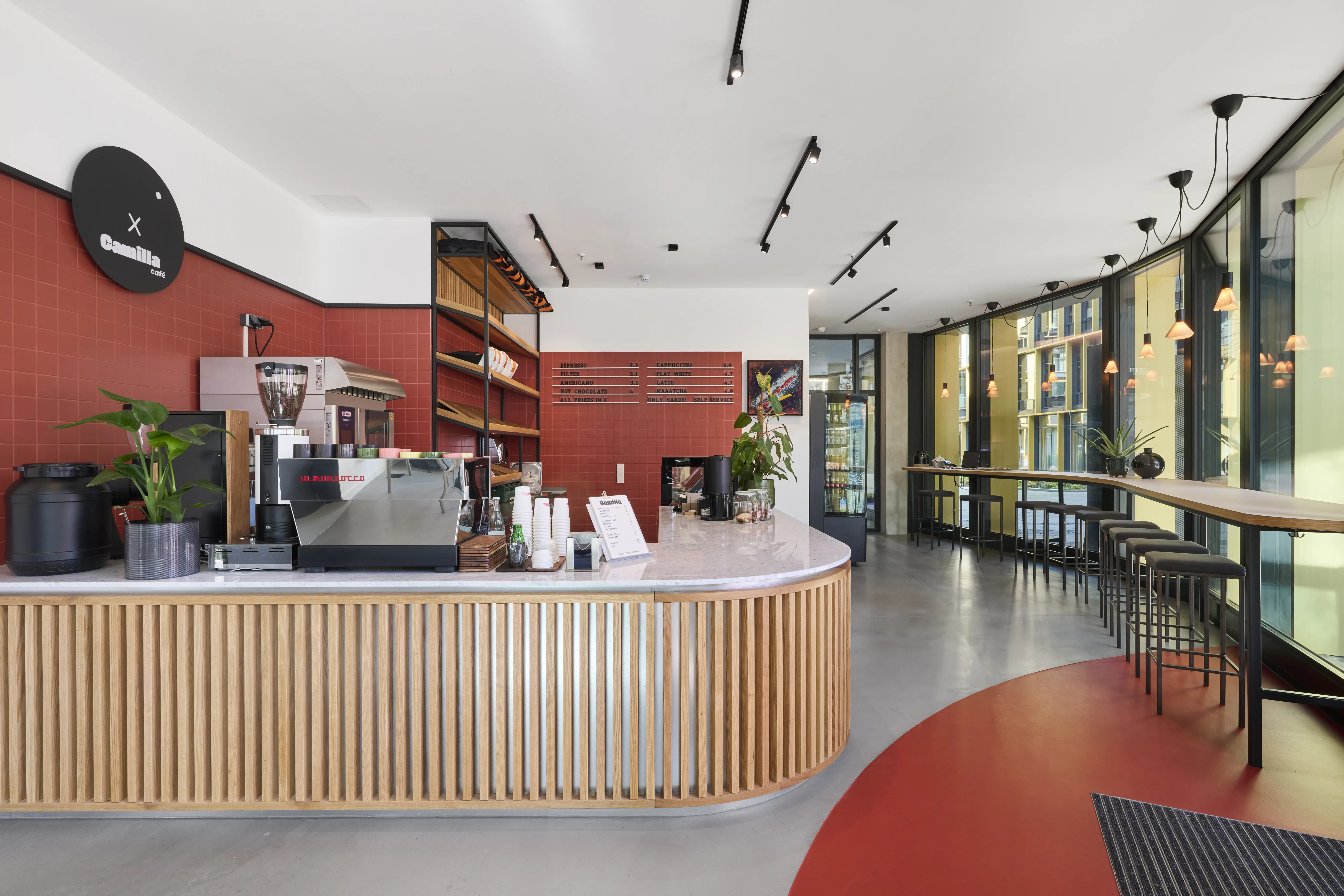 Interior of CAFÉ CAMILLA with a pastry display fridge on pink tiles, wooden shelf rack with products and terracotta-red tiled walls leading to a black door with PARKER BOWLES lettering.