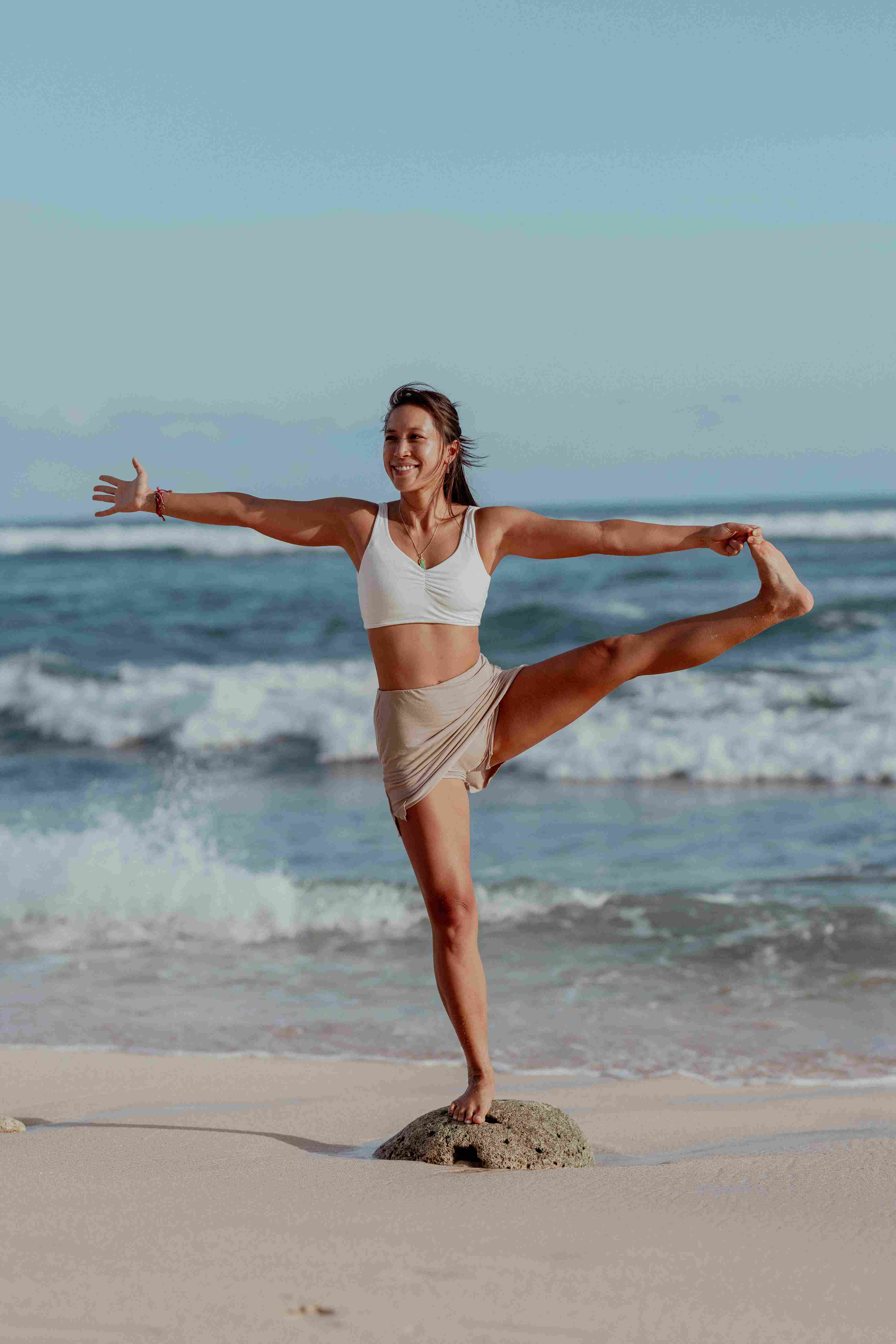 Woman practicing Utthita Hasta Padangusthasana balance pose on the white sand beach in Uluwatu.