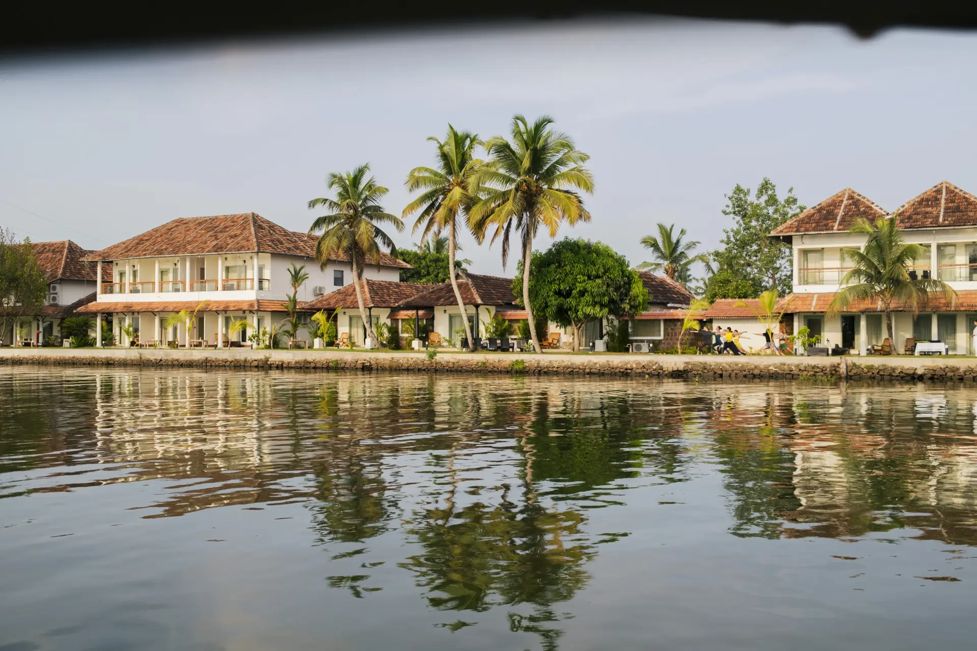 Panoramic view of Vedic Village Alleppey resort from the Kerala backwaters, guests practising yoga on the waterfront terrace with white heritage buildings and palm trees