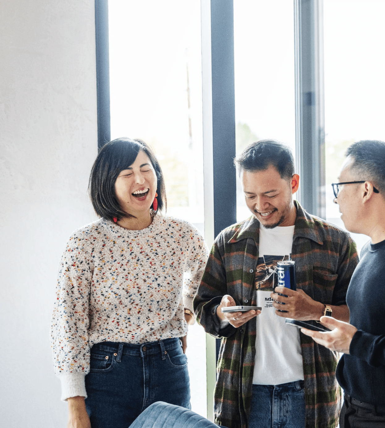 Three coworkers laughing together near large windows, looking at phones and holding a drink in a casual office setting.