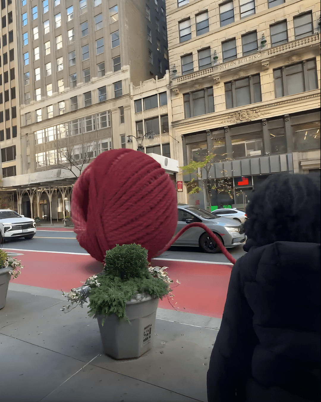 A massive red ball of yarn rolling down a busy city street past pedestrians and cars