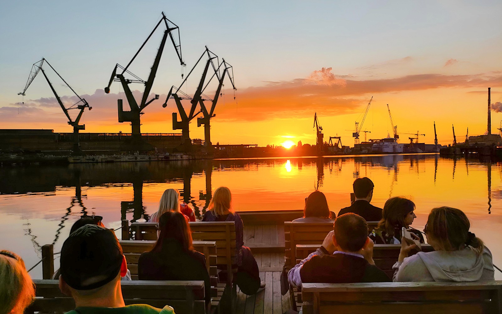 People on a river cruise in Gdańsk enjoying sunset with shipyard cranes in view.