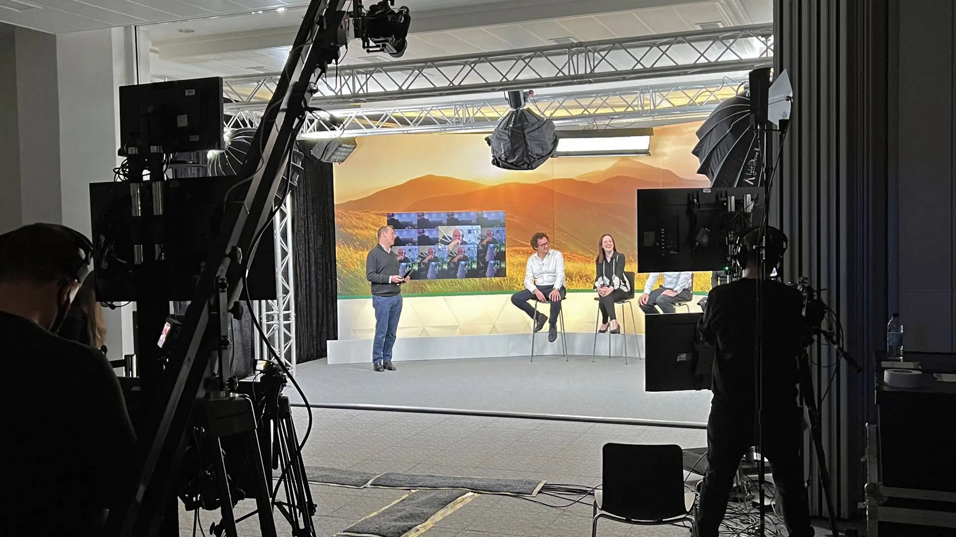 Wide shot of a live studio set with presenters seated on stage, lighting rigs, cameras, and crew operating during a broadcast.