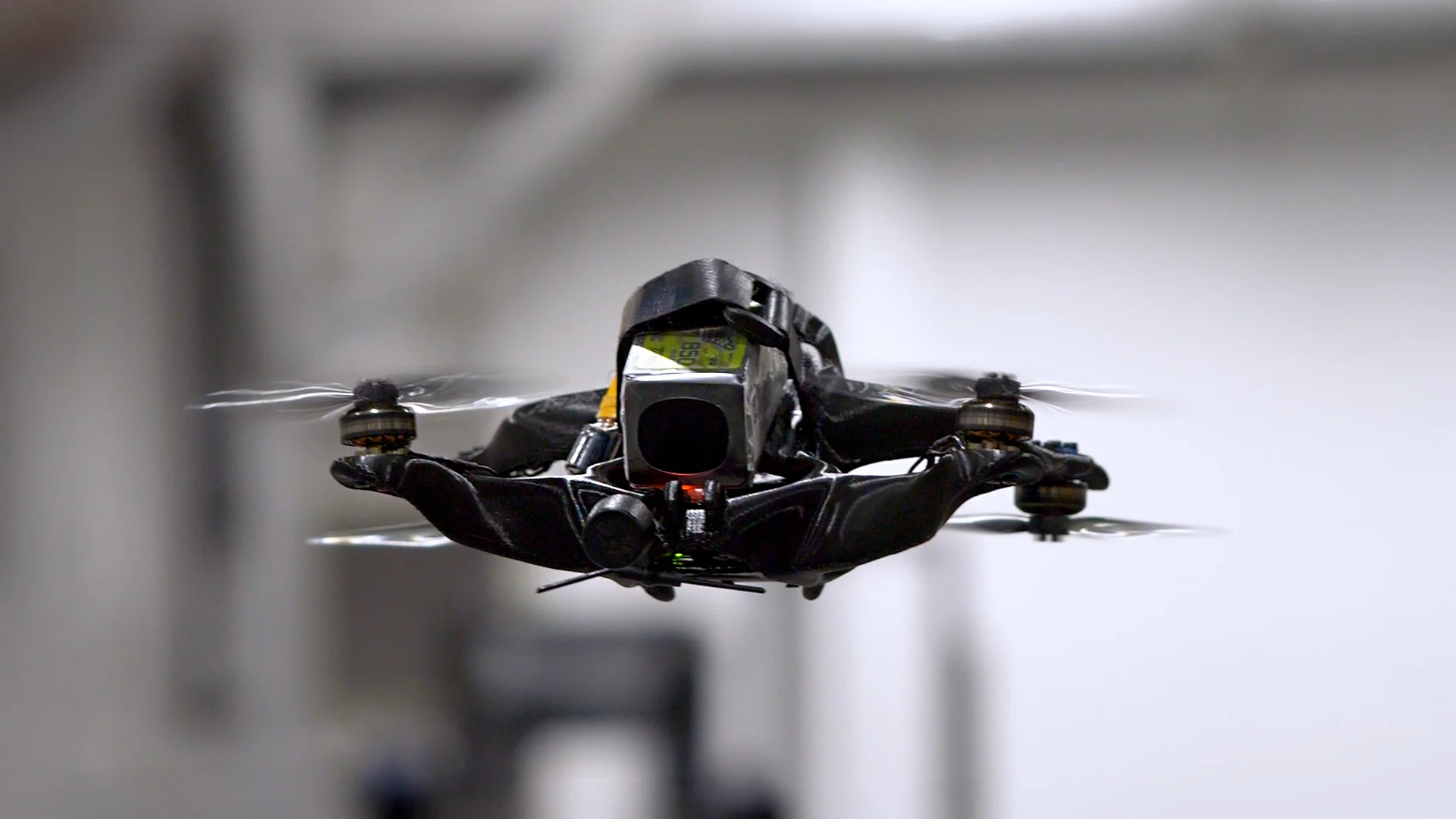 A sleek black drone hovers in mid-air, showcasing advanced quadcopter technology utilizing 3d printing against a blurred industrial background.