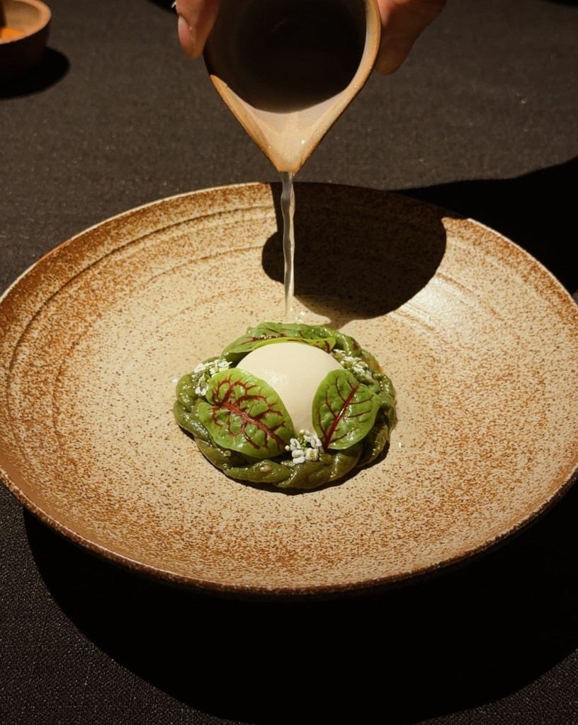 A beautifully plated dish featuring a creamy, green-hued element surrounded by decorative leafy greens and small white flowers, with a stream of sauce being poured over it.