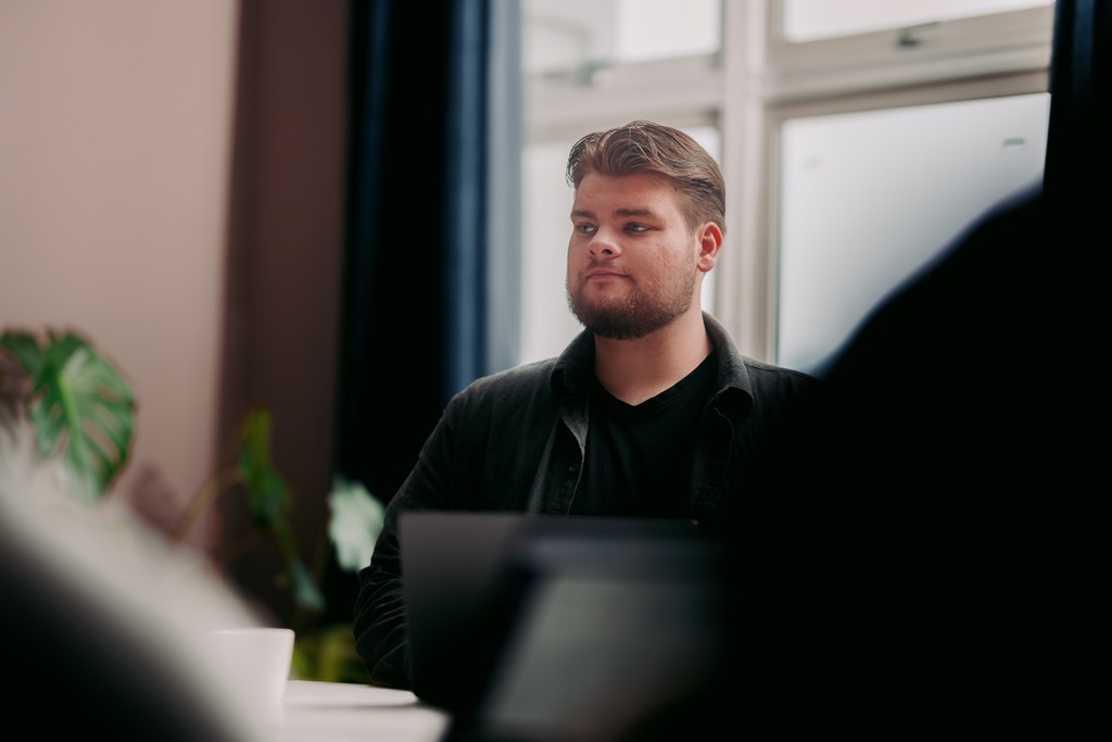In a modern office setting, a man with a beard is seated at a table, focusing intently, with a plant and large windows in the background.