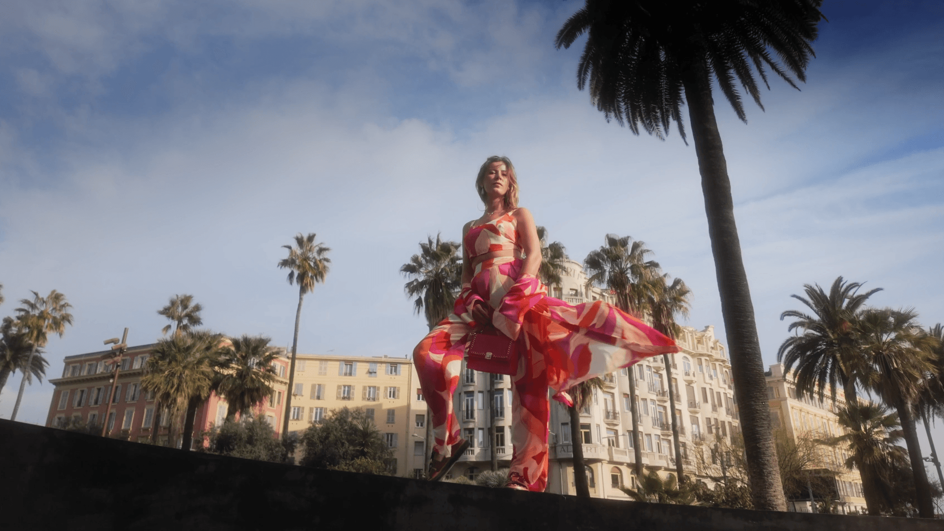 A person in a bright red outfit poses outdoors with palm trees in the background against a cloudy sky.