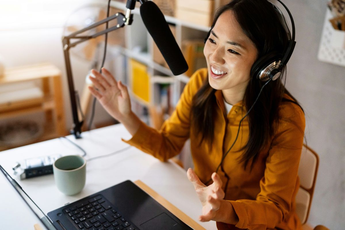 Young woman wearing headphones, enthusiastically speaking into a microphone while recording in a home studio with a laptop and a mug on the desk, representing podcasting, communication, and digital content creation.