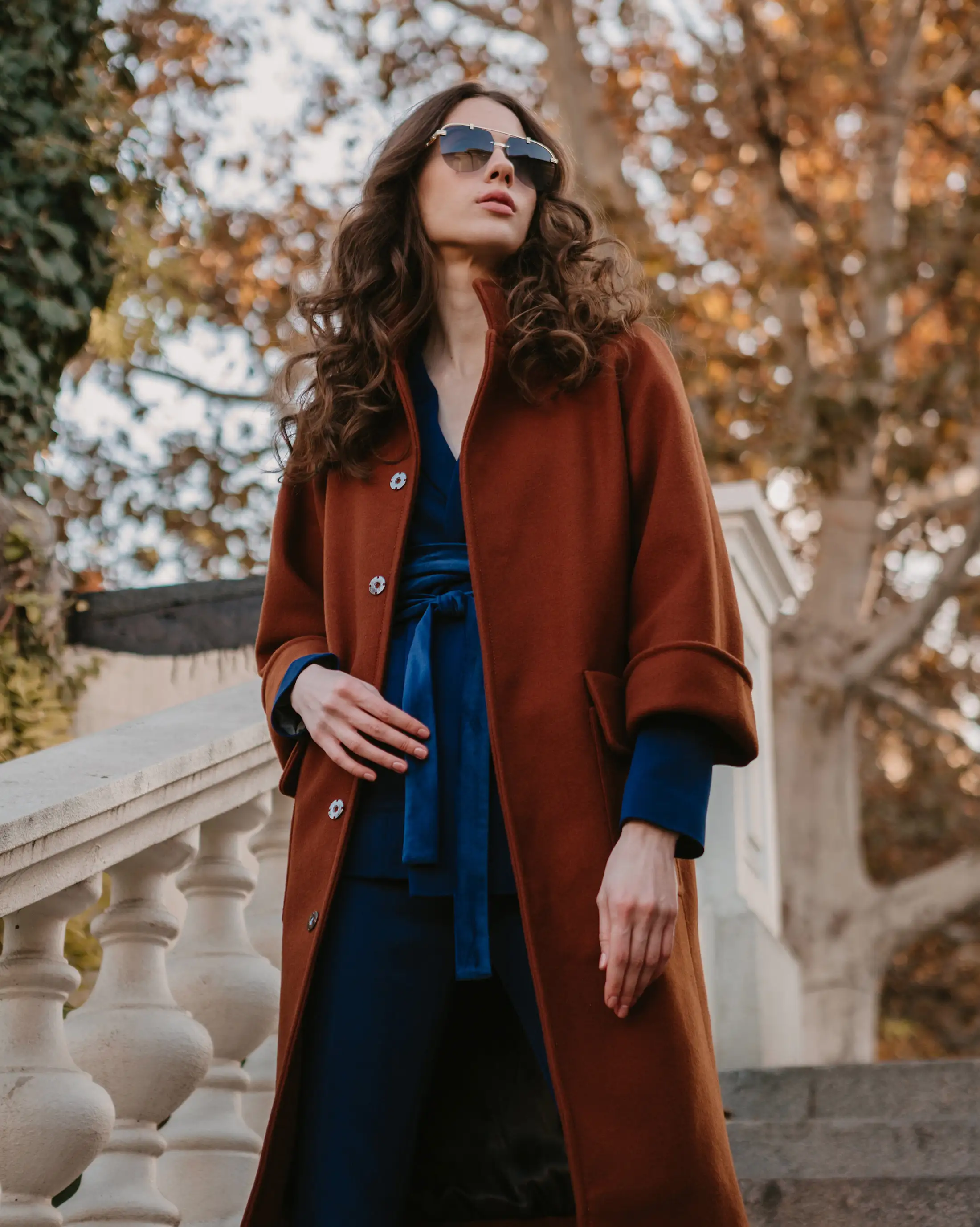 Model in a rust wool coat over a blue wrap suit on stone steps beneath autumn trees