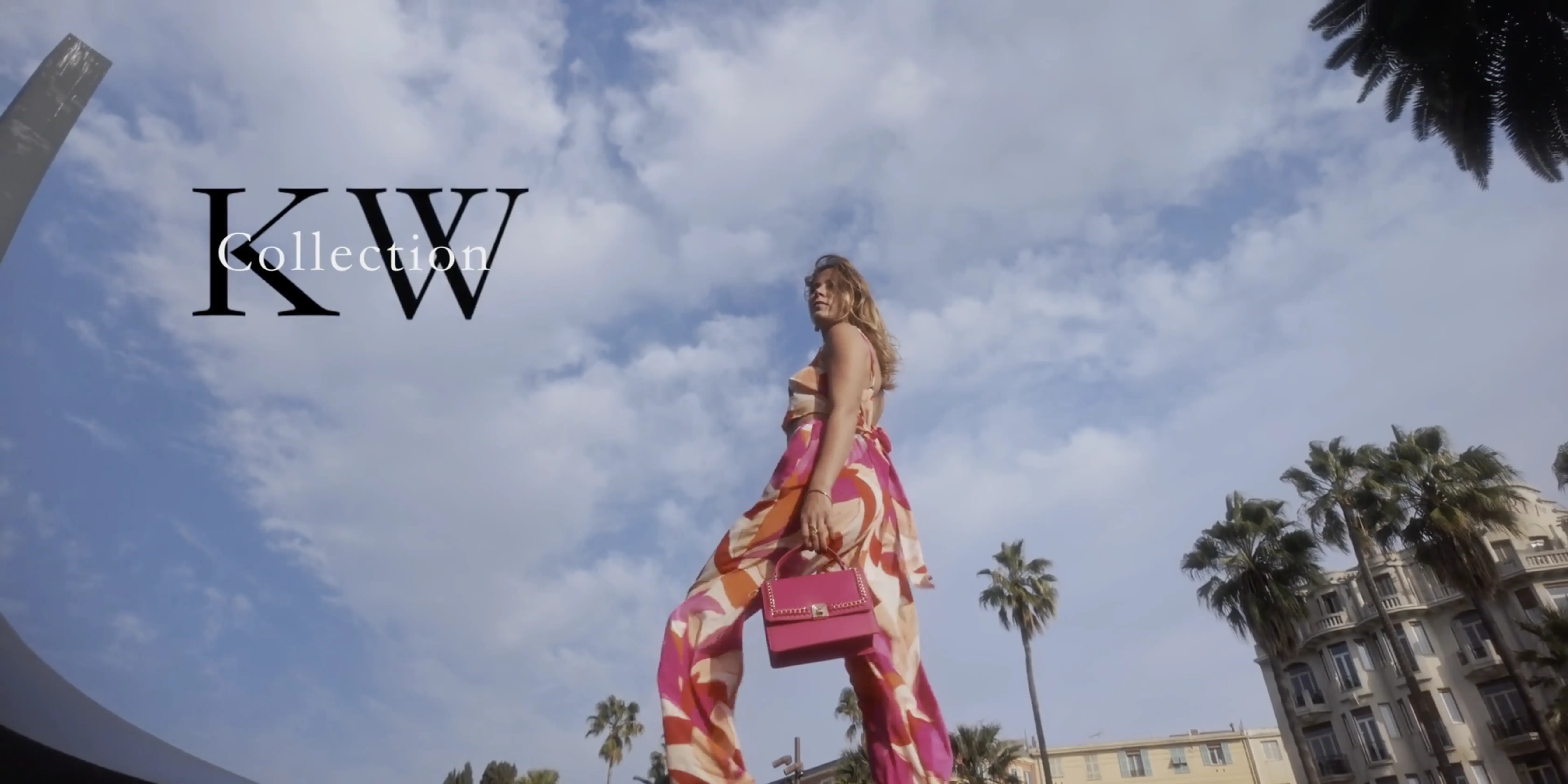 A woman dressed in vibrant, flowing patterned pants and a matching top holds a pink handbag, standing confidently against a backdrop of palm trees and historic buildings under a partly cloudy sky, with the text "KW Collection" displayed prominently.