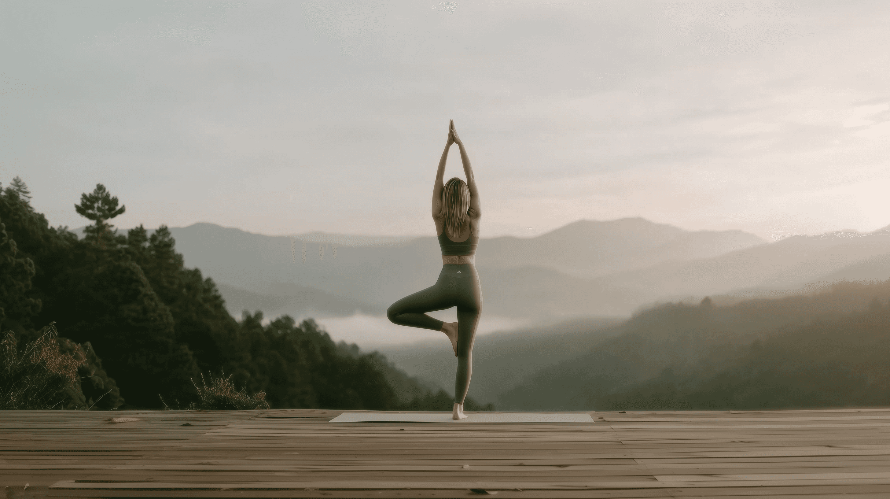 Woman practicing yoga in tree pose on a wooden deck overlooking misty mountain landscape at sunrise.