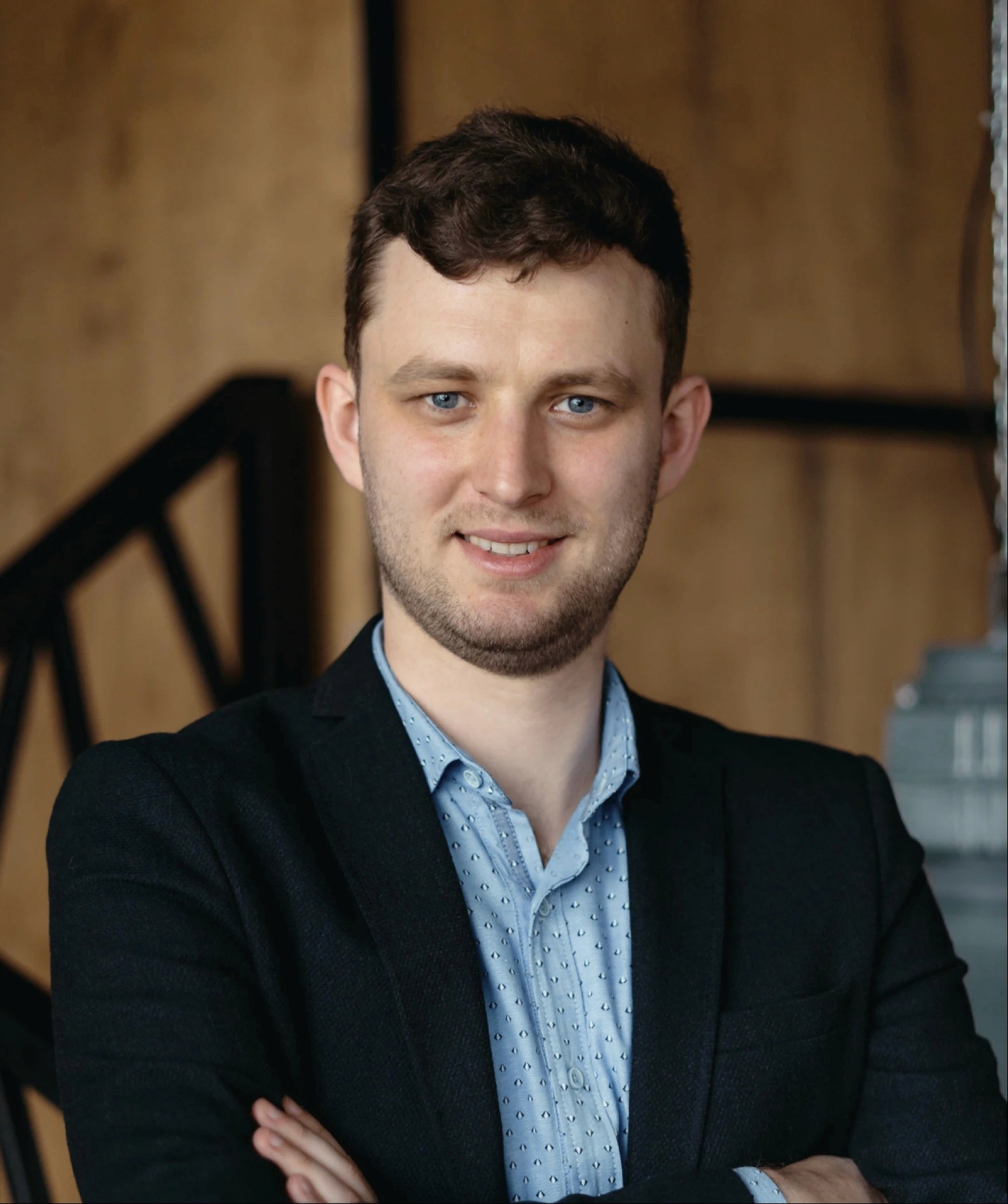 A young man with short dark hair smiles confidently, wearing a suit jacket, against a rustic wooden backdrop.