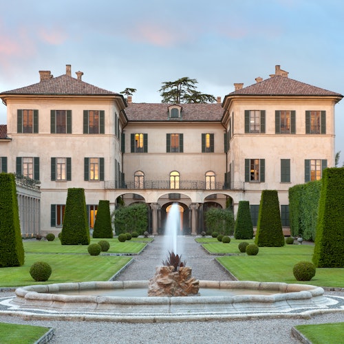 Stor, elegant herrgård med symmetriska torn, omgiven av välskötta trädgårdar, formklippta träd och en central fontän.