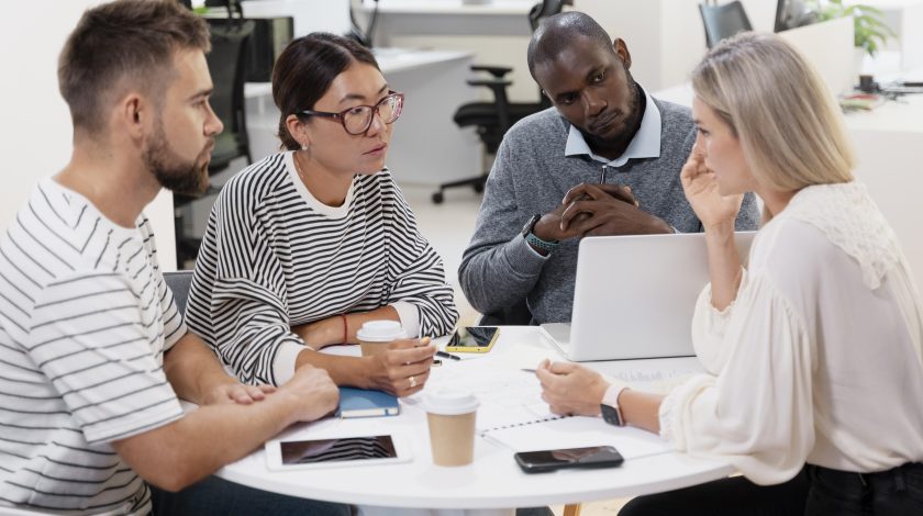 Four diverse colleagues engage in a collaborative discussion around a white table in a modern office, with notebooks, a laptop, and coffee cups visible.