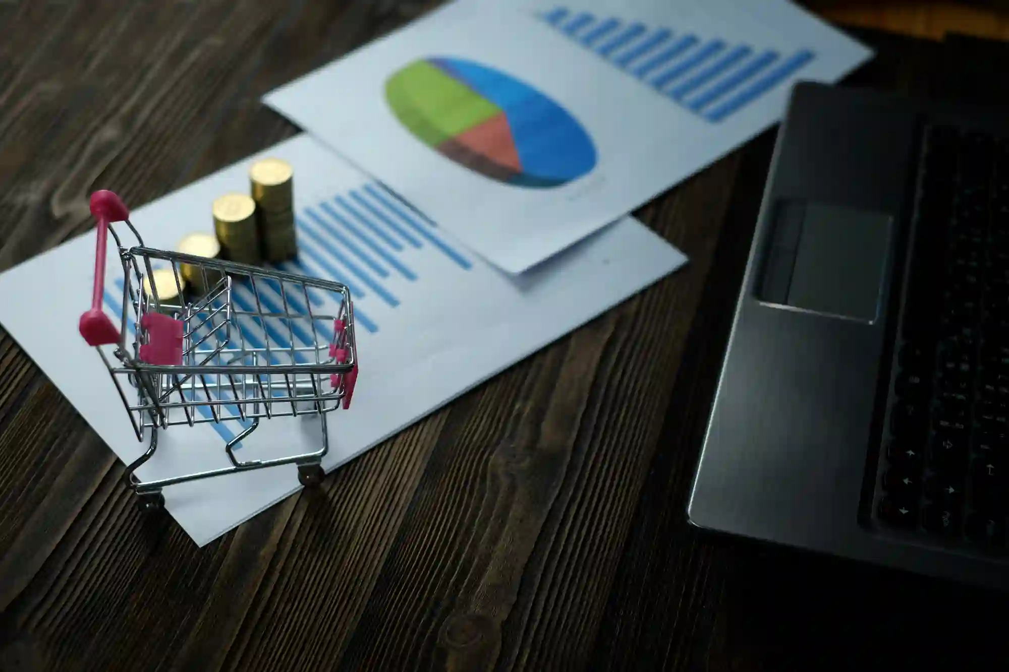 Miniature shopping cart with coins, laptop, and financial charts, suggesting e-commerce sales analysis or online business growth.