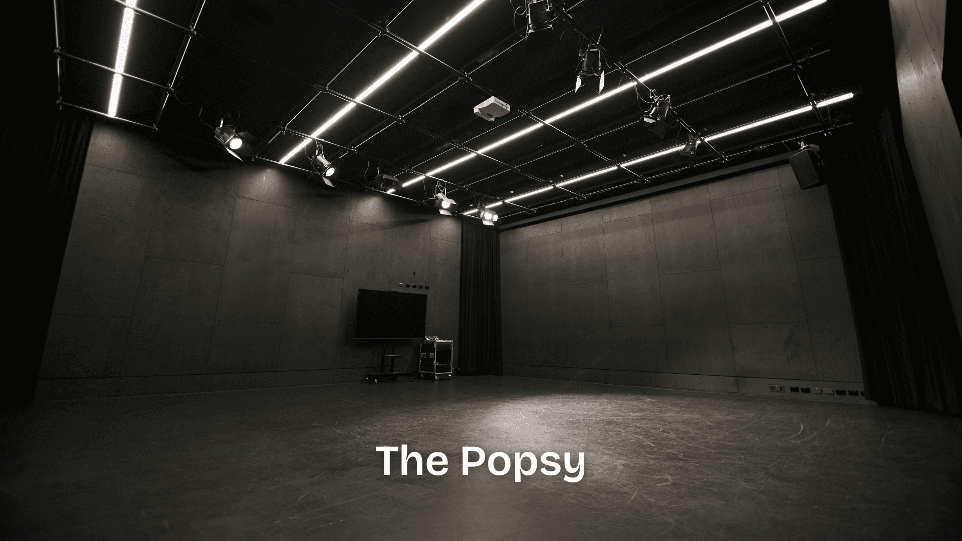 A black-walled studio theatre with a lighting grid and movable AV equipment, designed for small-scale performances, rehearsals, and development showings.