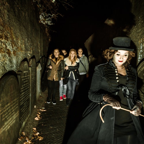 빈티지 의상을 입은 여성이 무덤이 늘어선 어두운 좁은 복도를 통해 그룹을 안내하고 있습니다. 그룹은 관심을 보이는 것 같습니다.