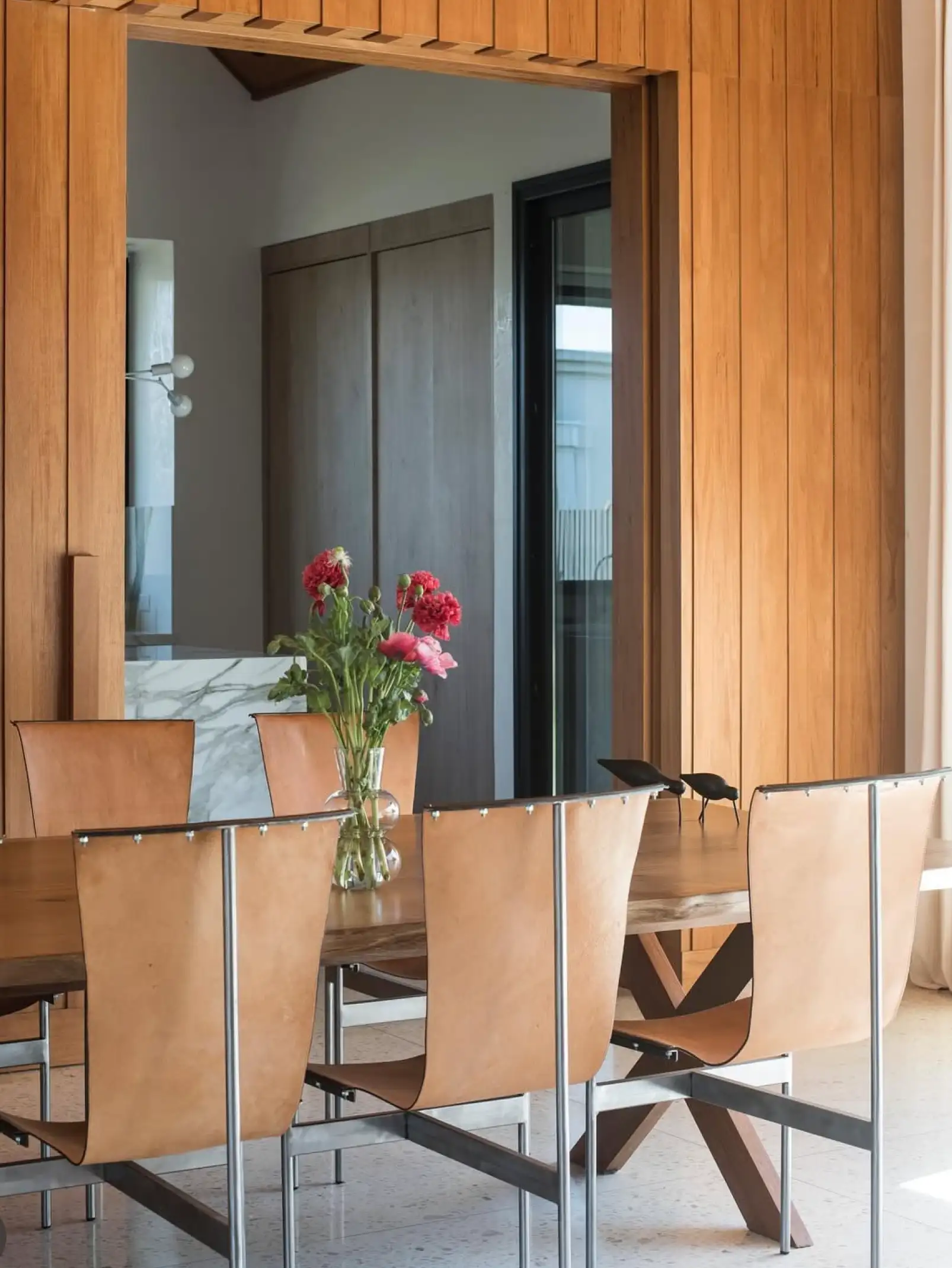 Comedor con sillas de cuero y flores rojas sobre mesa de madera para diseñadores que valoran materiales cálidos y detalles naturales.