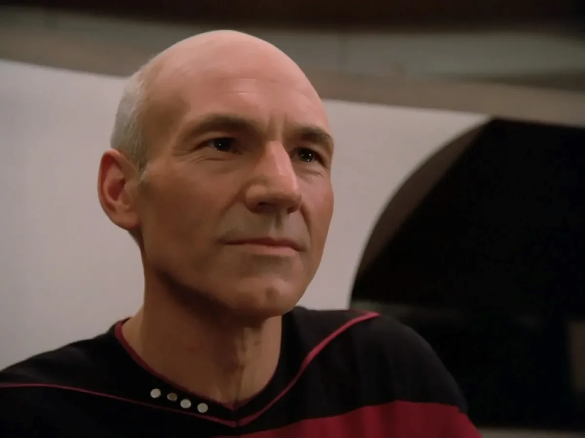 A bald man in a futuristic uniform sits in what appears to be the interior of a spaceship, with muted lighting highlighting his thoughtful expression.