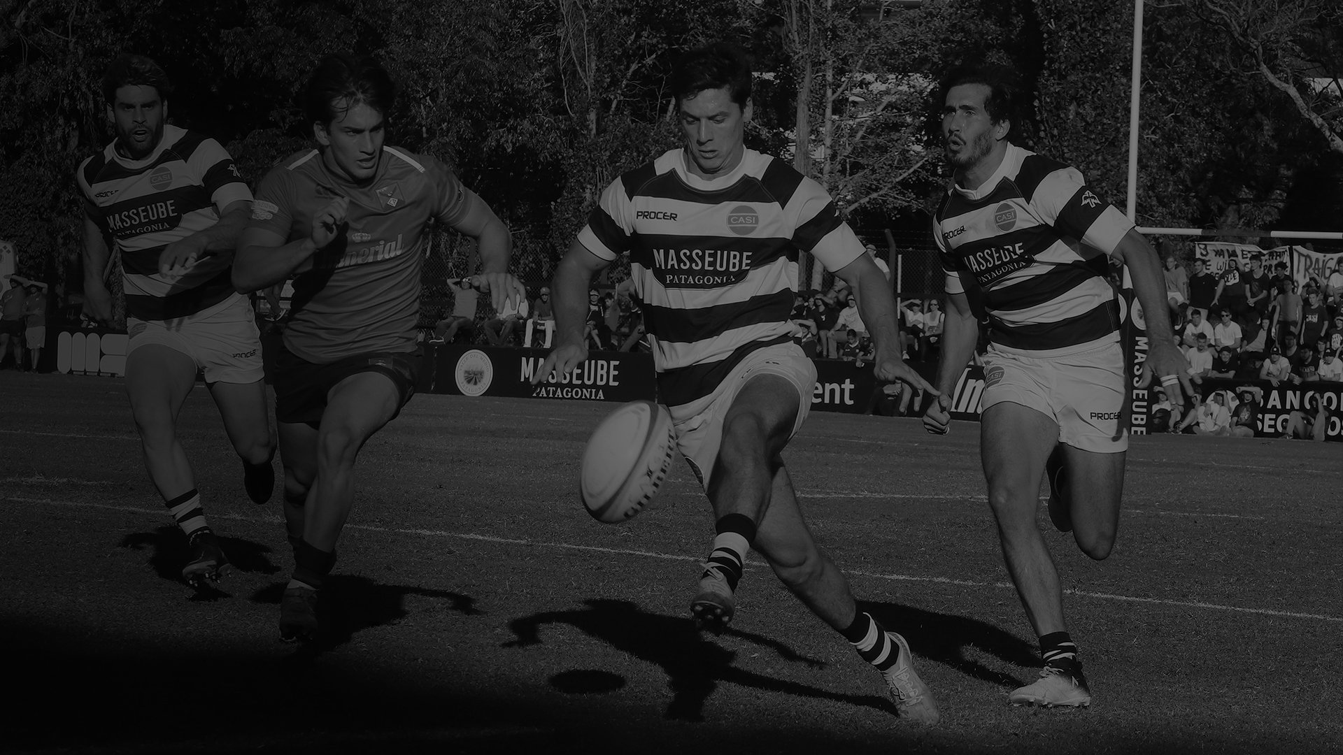 Rugby players intensely sprint toward a rugby ball on a grassy field. One player prepares to kick as focused teammates and an audience watch.