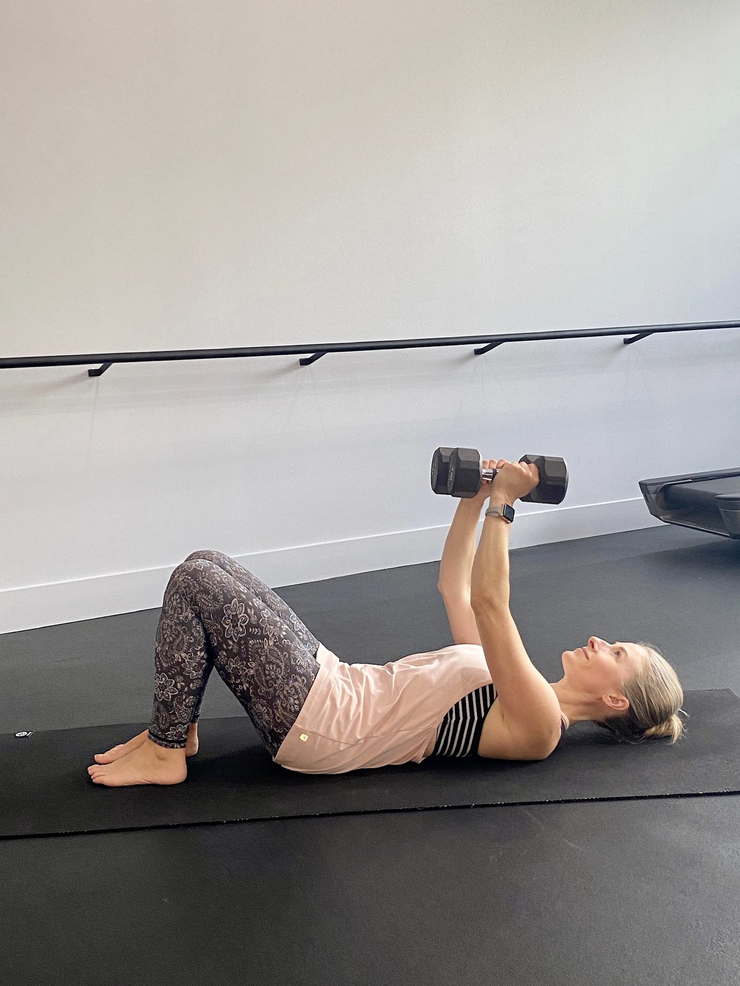 A woman is lying on a black exercise mat in a gym, performing a chest press with dumbbells, dressed in a pink tank top and patterned leggings.