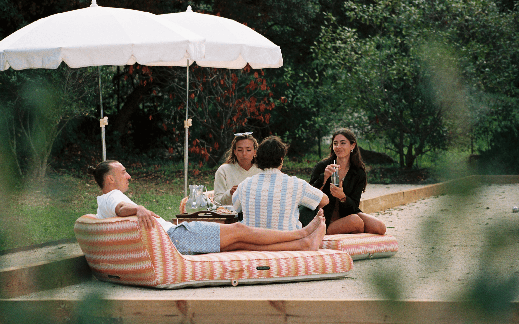 Friends lounging on a sunset-coloured chevron pool float under a white parasol on a garden path.